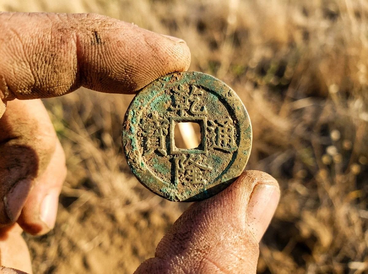 Weathered Chinese cash coin with square center hole held in dirt-covered fingers above dry grass